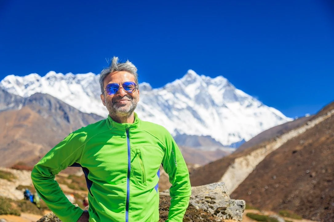 Ashok hiking in front of snow-covered mountains, wearing a bright green jacket.